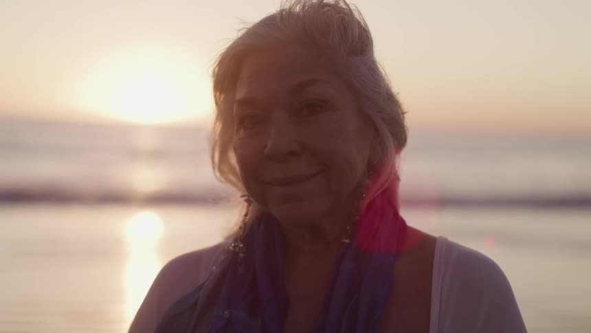 Elderly woman standing on the shoreline of a beach at sunset looks at the camera