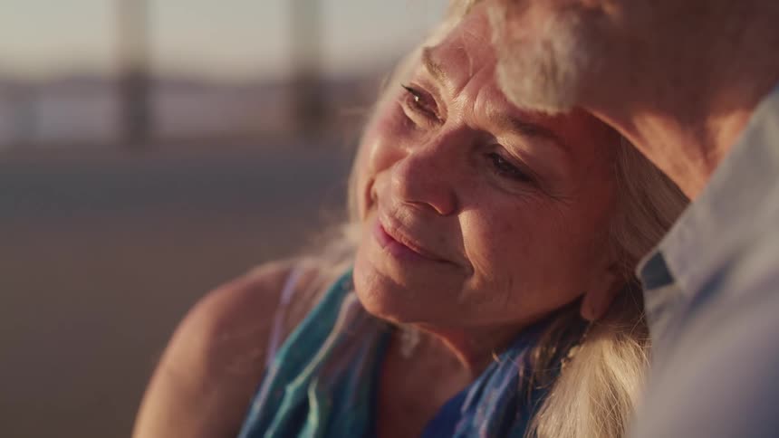 Elderly woman and man hold each other and watch sunset on the beach