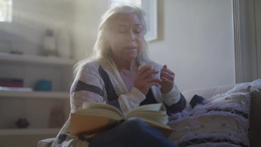 An elderly woman drinking coffee while reading a book at home