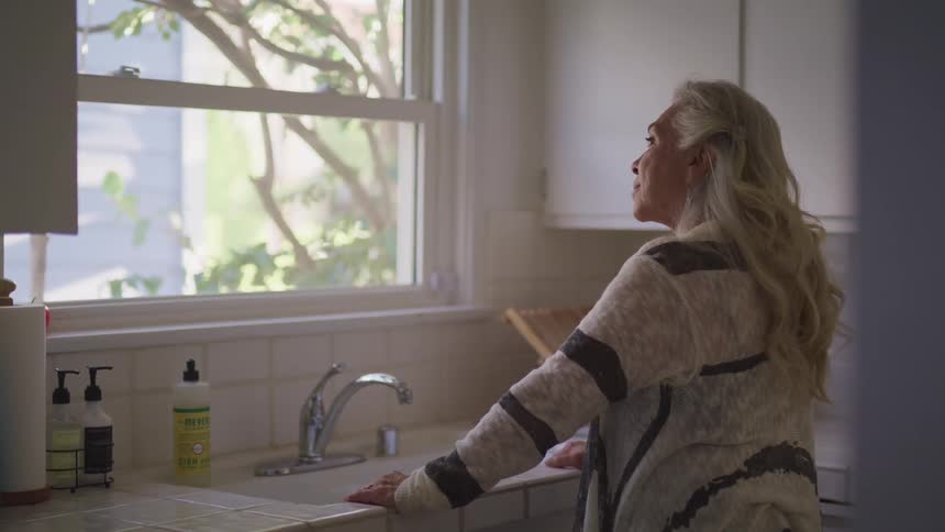 Elderly woman with earrings and grey hair looks out the kitchen window