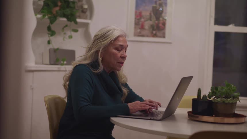 Elderly woman with grey hair and a sweater sits at a table and uses a macbook pro