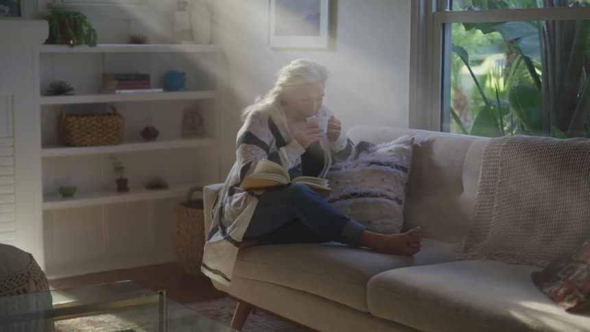 A female caucasian laying on a couch drinking and reading from a book