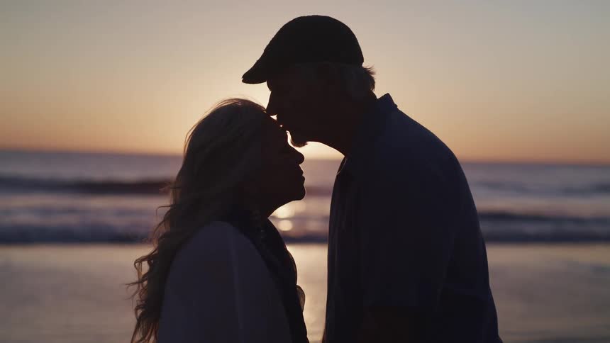Husband and wife stand on the beach at sunset facing one another as husband leans in and kisses his wife on the forehead