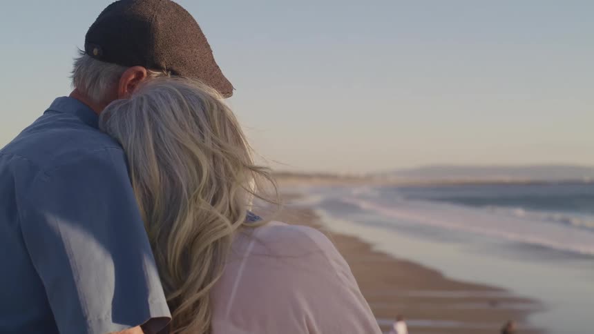 Elderly man with a blue shirt and brown tweed cap holds elderly woman with grey hair on the beach next to the sea
