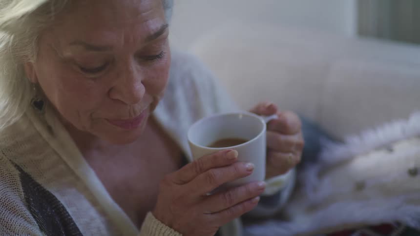 Elderly woman with grey hair sitting on a couch drinks a cup of tea