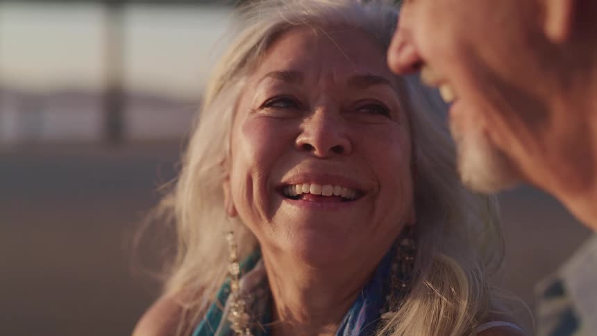An elderly woman smiles as she looks upon her male companions face before resting her head on his shoulder