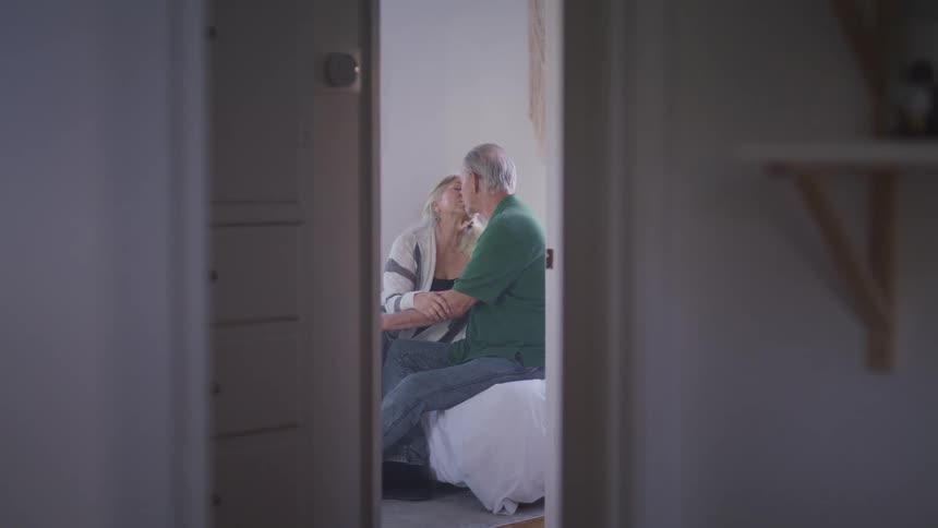 Elderly man with a green polo shirt kisses elderly woman with grey hair on the bed