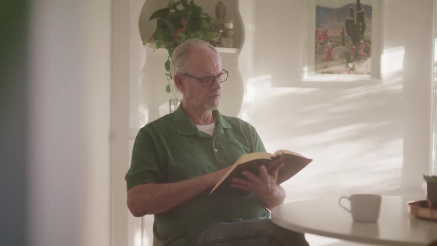 An elderly man sits at his kitchen table with a cup of coffee and reads a book