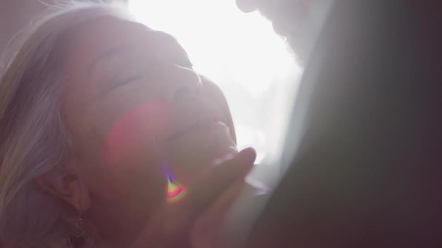 Elderly man kisses elderly womans cheek and they look into each others eyes with love as the shines through windows