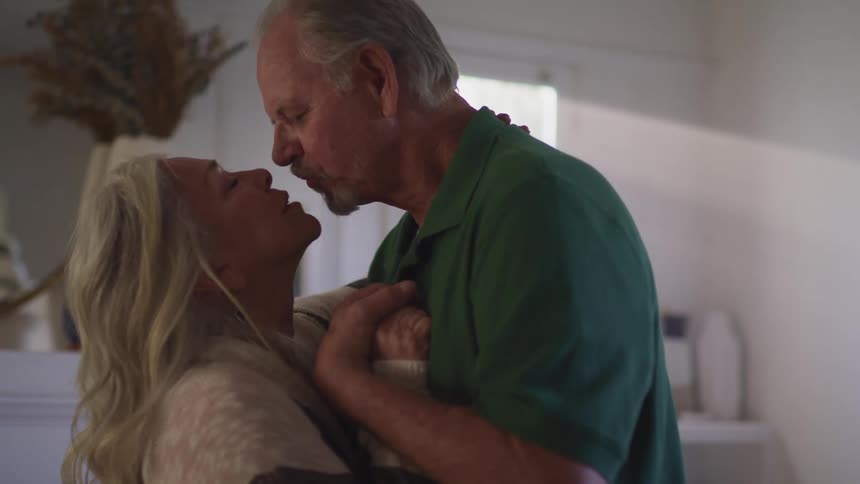 An elderly man and woman share a kiss indoors as sunlight shines in through a window behind them