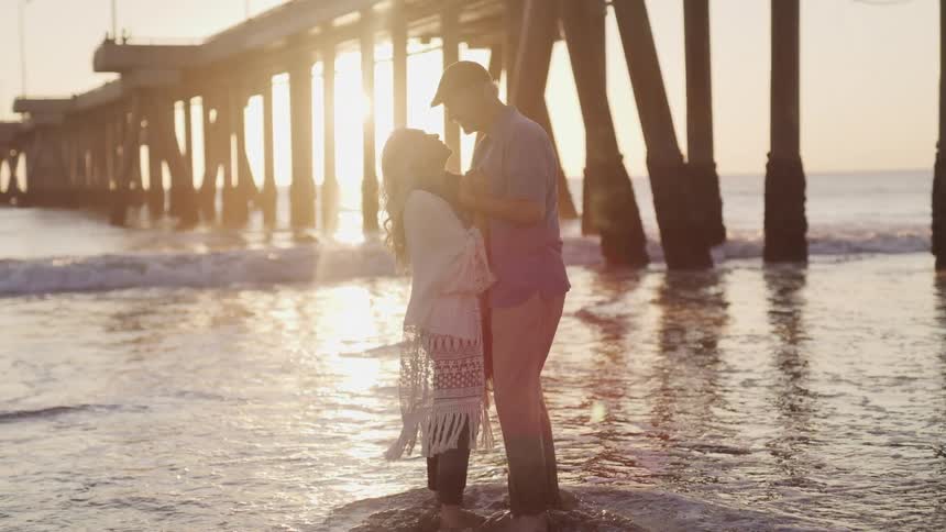 An elderly couple embrace and gaze at one another while standing in the low water under a boardwalk at the beach at sunset