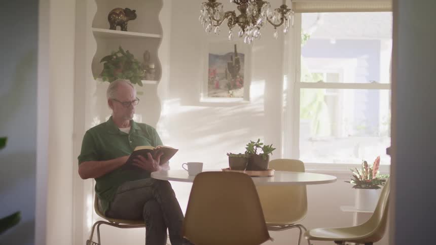 An elderly man sitting in a kitchen reading from a book