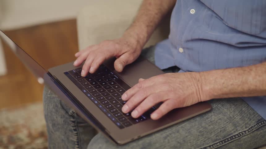 A man sits with a laptop on his lap and types on it