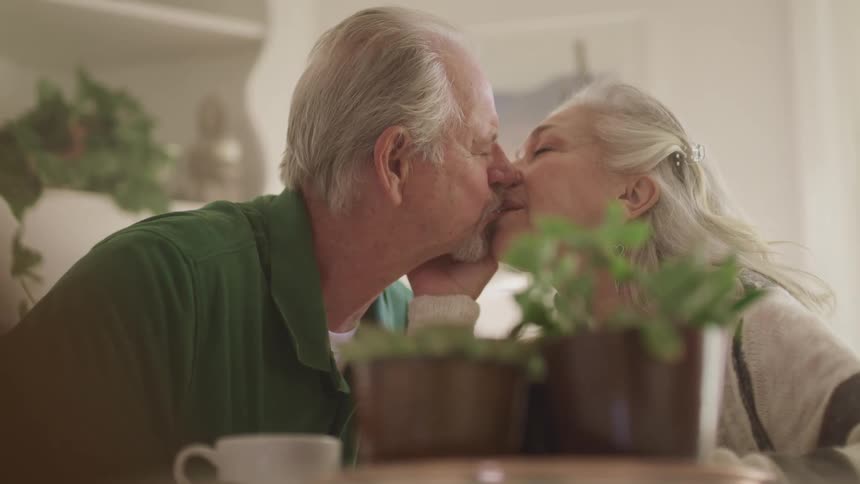 An elderly couple share a kiss while sitting at a coffee table holding potted plants