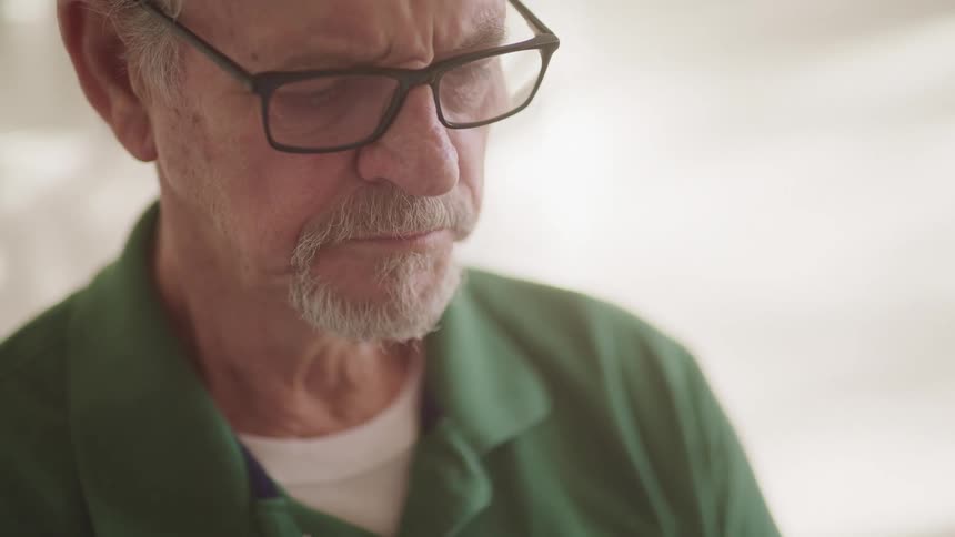 Elderly man with a goatee beard a green polo shirt and glasses looks down