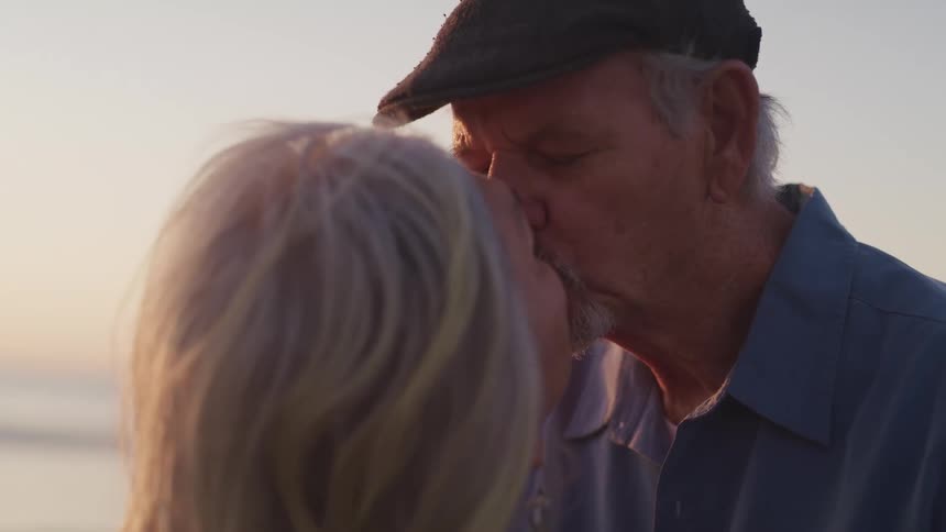An elderly couple share a kiss on the beach at sunset