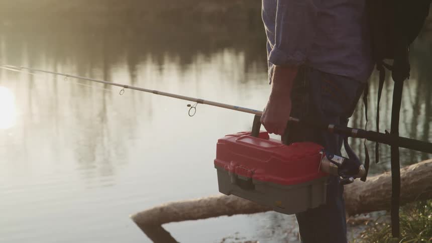 Fisherman stands by a lake while holding a fishing pole and tackle box