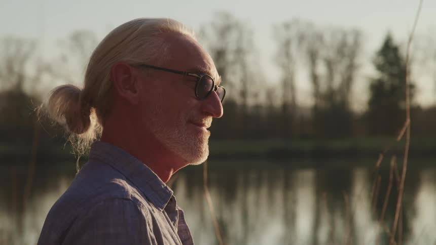 Senior wearing sunglasses and a pony tail stands near a lake at sunset