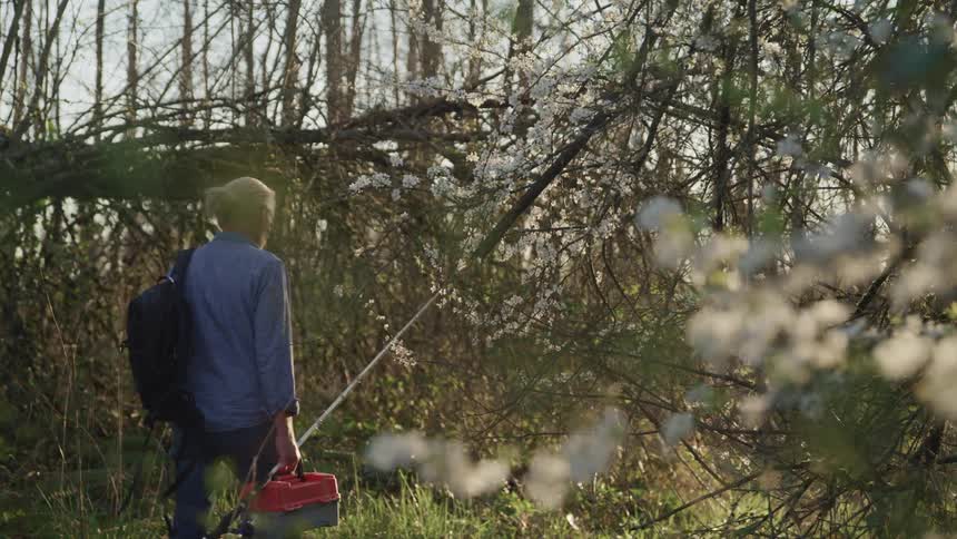 A man walking through a forest carrying a fishing box and fishing rod