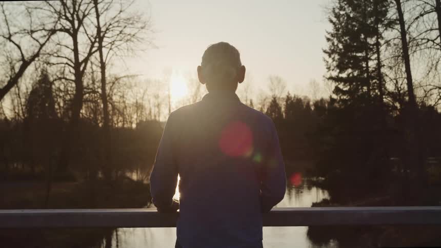 A person standing on a bridge looks out over the small river running beneath it on a cloudy evening