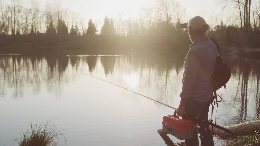An old man standing on a lakeshore while carrying fishing gear shields his eyes from the sun as he looks out over the lake