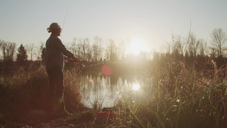 Elderly man with glasses and long hair casts a fishing pole in the lake at sunset