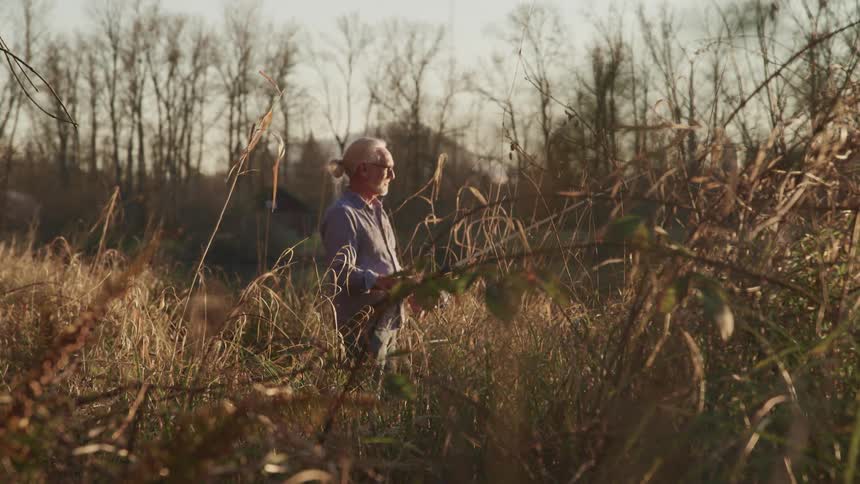 Man stands with fishing pole in field of dried grasses near leafless trees
