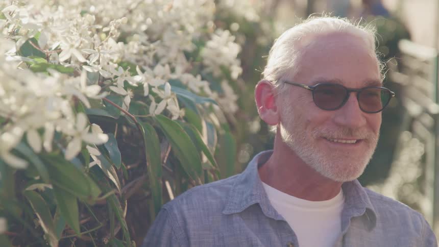 A man wearing sunglasses smiles while standing next to flowering shrubs