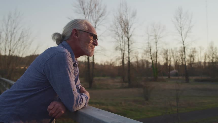 A man with glasses leaning on hand railing with trees and the sky in the background