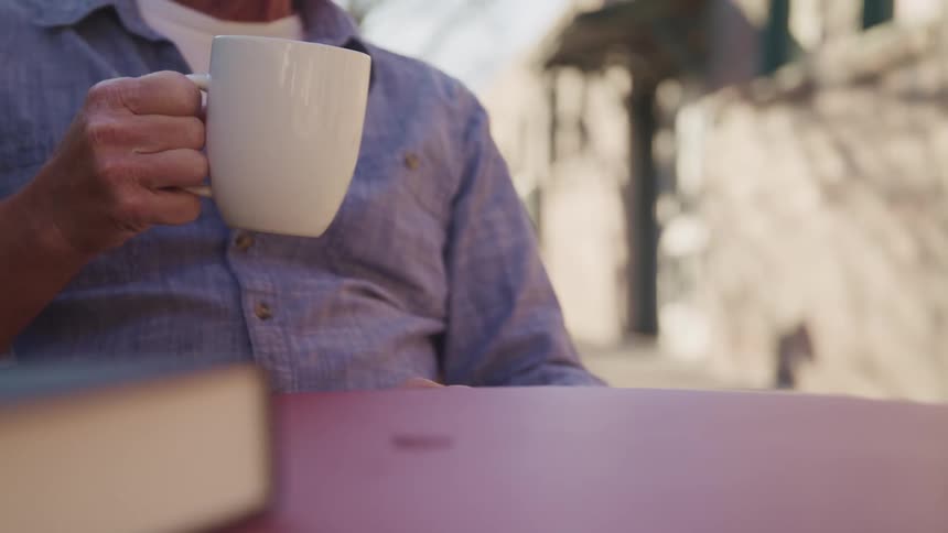 A man drinks a hot beverage from a white ceramic mug sitting in the table of an outdoors cafe