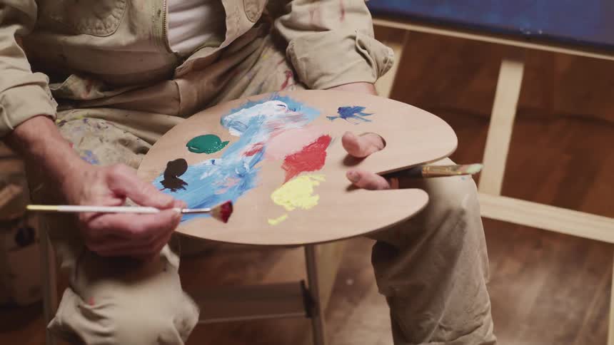 A painter holds a palette covered in paint as they sit on a stool in front of an easel holding a painting