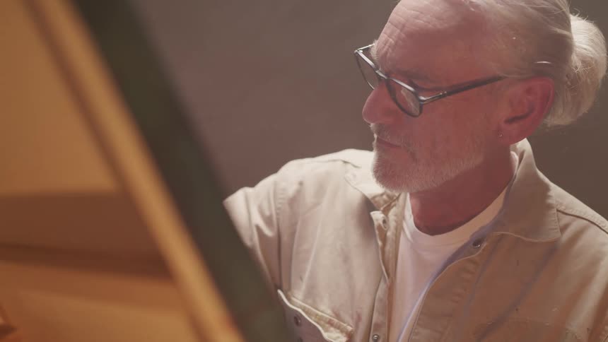 An older male painter with a short ponytail and glasses applies paint to a canvas