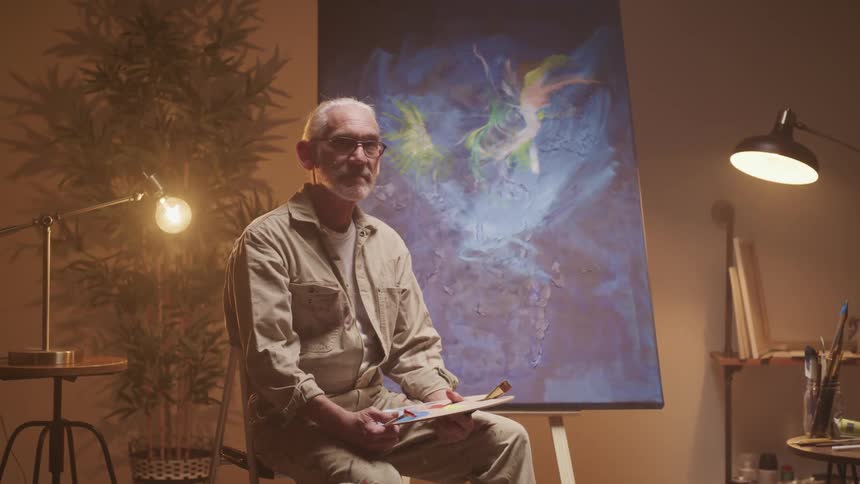 An artist sits and holds his palette in front of a large painting