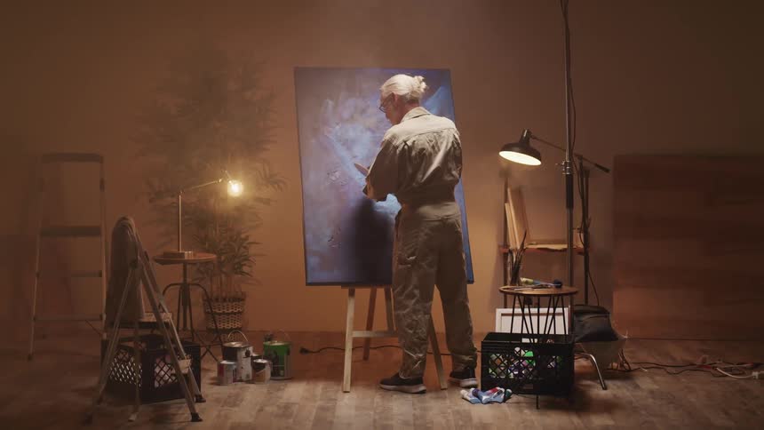 An elderly man stands in front of a canvas on an easel and uses paint from a palette to complete a painting