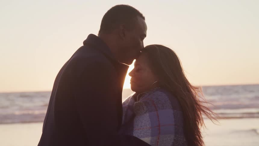 A woman grimaces after her boyfriend kisses her on the forehead as they stand on a beach next to the ocean
