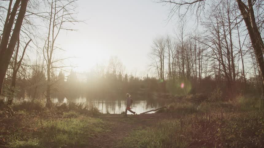 An elderly fisherman carries his fishing equipment as he walks along the shore of a lake in the morning