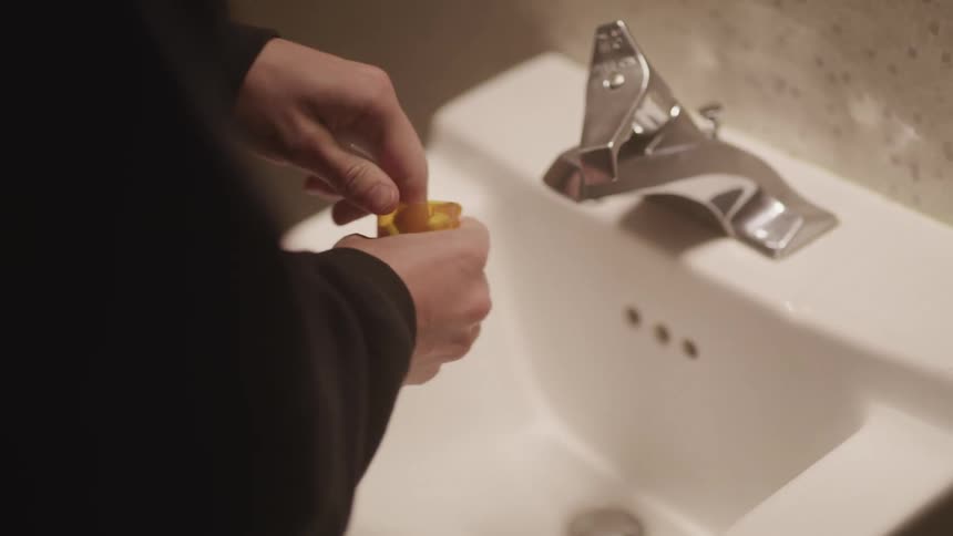 A female standing over a sink opens a bottle of pills and grabs some out as others fall in the sink