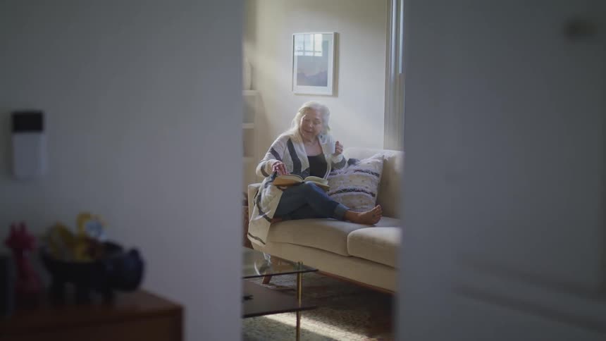Elderly woman sitting on a beige sofa reads a book in the living room