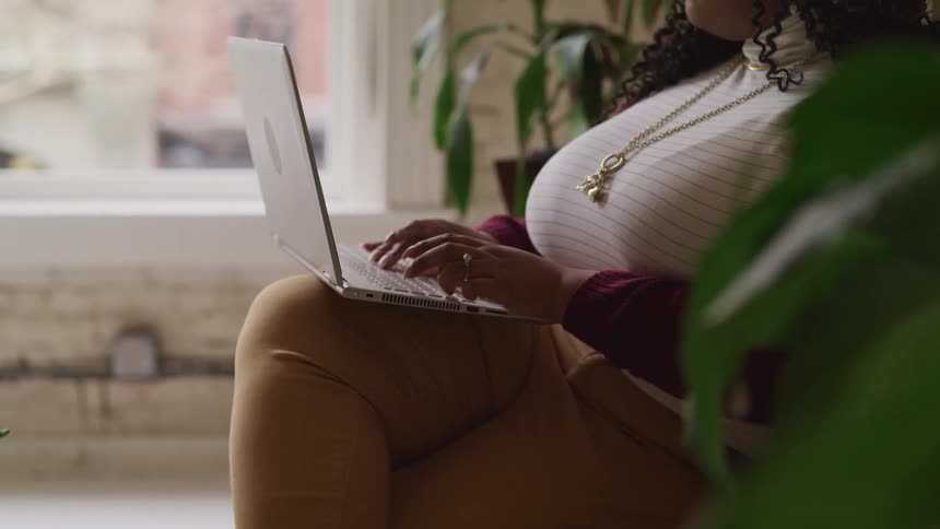 Black chubby woman with curly hair sits on a sofa and uses a grey laptop in a room