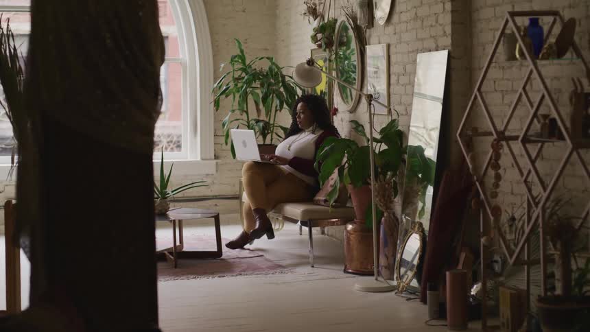 A woman sitting in a room typing on a laptop surrounded by plants with a window in the background