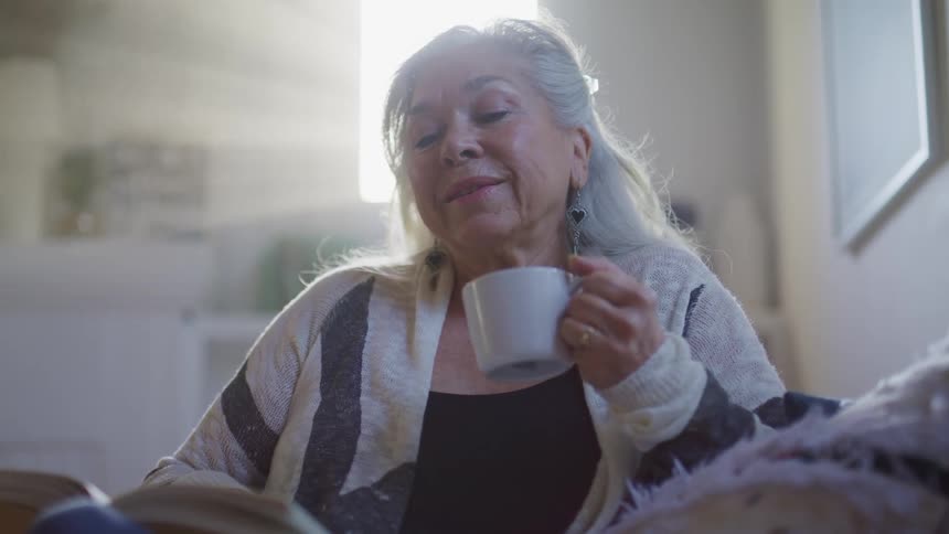 An elderly woman sits on a chair holding a small mug and turns the page of a book