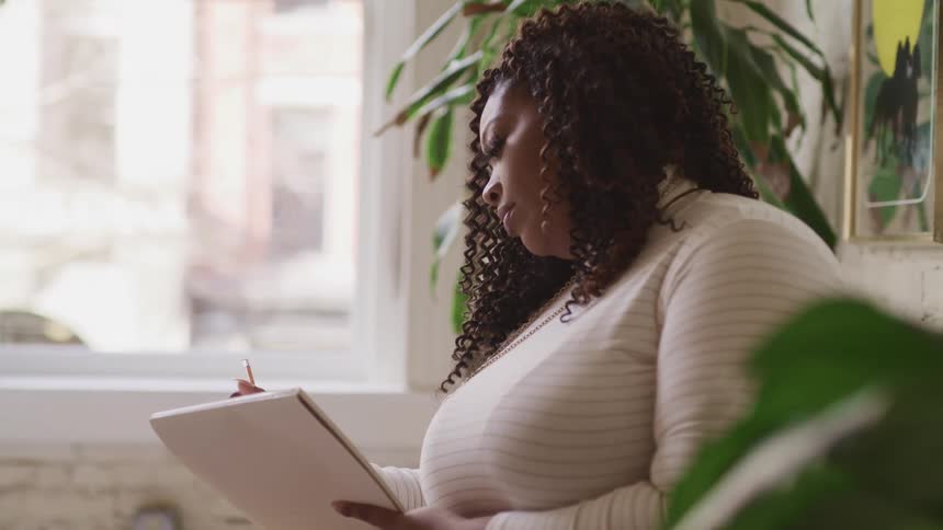 Chubby black woman with curly hair sits on a sofa and writes things down in a notebook