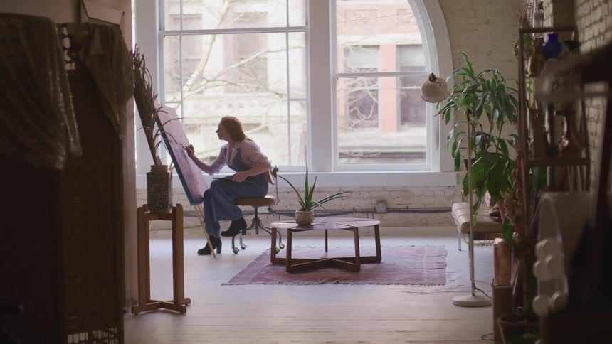 An artist paints on a large canvas in front of a living room window