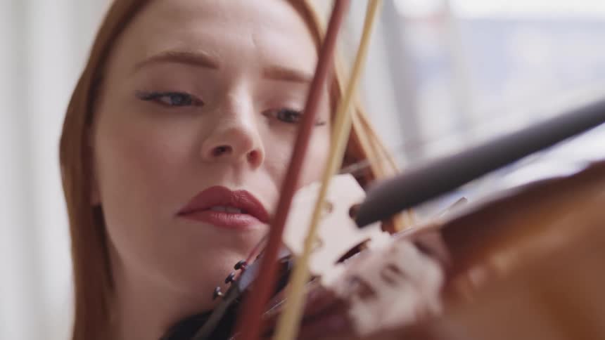 Woman with red hair and fair skin holds violin under her chin and play with a bow
