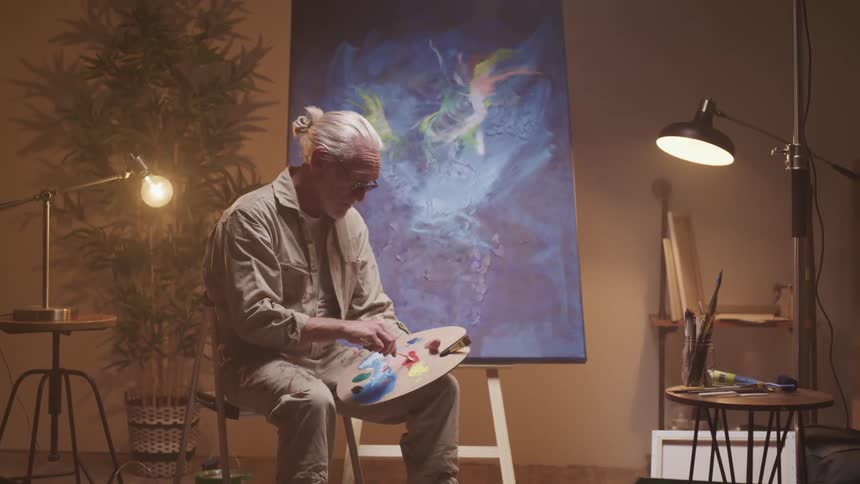 An elderly painter mixes paint on a palette while sitting on a stool in front of a paint on a large canvas