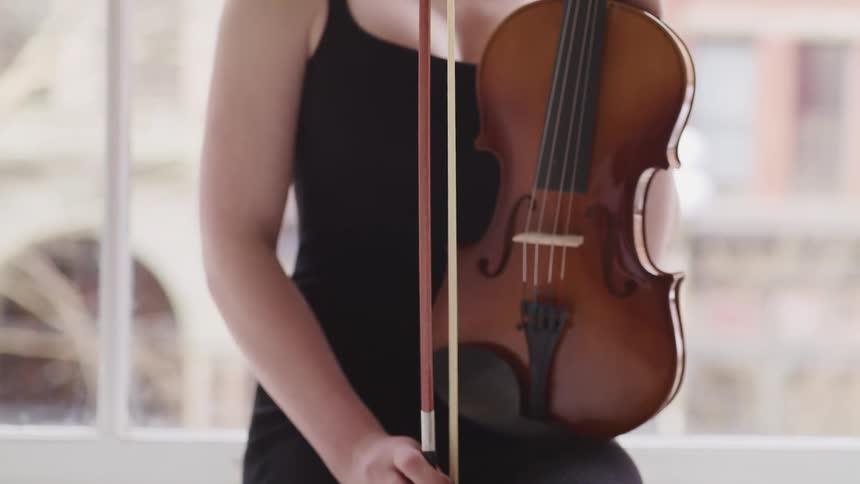 Woman with a fair skin a black dress sits on a windowsill and hold a violin with a bow