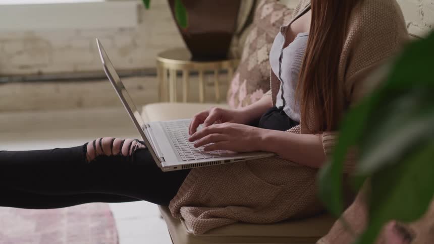 Woman with a ripped jeans sits on a sofa and types on a laptop keyboard