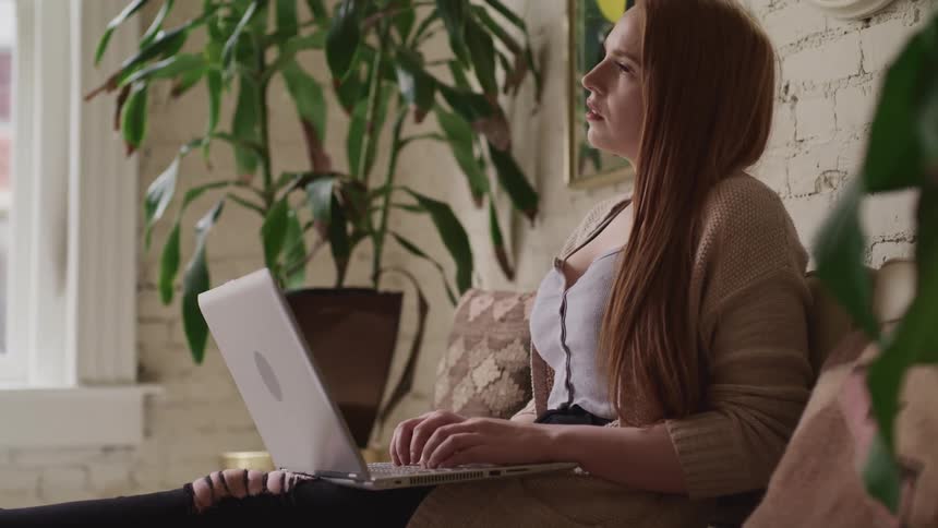 Woman sitting near painted brick wall bordered by houseplants looks into space while her hands rest on laptop keyboard