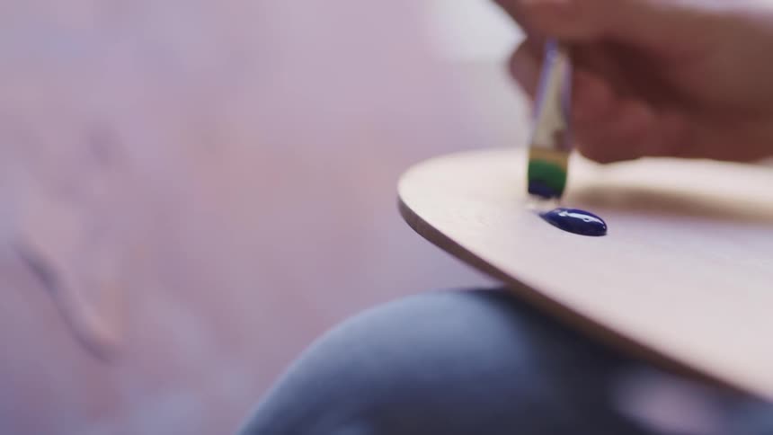 A persons hand holding a paint brush dipping it in blue paint on a painting pallet