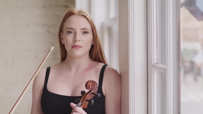 Young woman holding a violin next to a window looks at the camera
